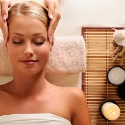 Blond kobieta z zamkniętymi oczami relaksuje się podczas masażu głowy; obok leży ręcznik i bambusowa mata z dekoracjami i kosmetykami.