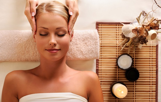 Blond kobieta z zamkniętymi oczami relaksuje się podczas masażu głowy; obok leży ręcznik i bambusowa mata z dekoracjami i kosmetykami.
