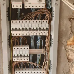 Rozdzielnica elektryczna z zamontowanymi bezpiecznikami Legrand, widoczne kable różnego koloru i przekroju, zamocowane w listwach zaciskowych, ściana w tle z surowym tynkiem.