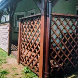 Drewniana pergola z kratownicą, brązowy słupek z rynną, widok z zewnątrz na tle zielonej ściany budynku z oknami.