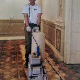 Mężczyzna w okularach cyklinuje parkiet maszyną w pomieszczeniu z dekoracyjnymi ścianami i oświetleniem.