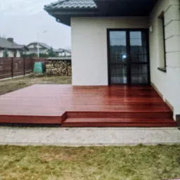 Nowoczesny taras drewniany z egzotycznego drewna, przylegający do budynku z czarnymi drzwiami balkonowymi, widok na trawnik i drewniane ogrodzenie w tle.