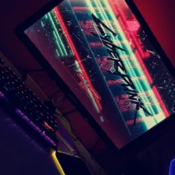 Monitor wyświetlający ekran gry Cyberpunk 2077, podświetlana klawiatura i podkładka pod mysz z motywem pająka w ciemnym pomieszczeniu.