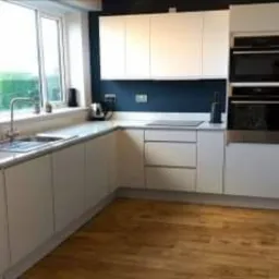 Jasna, nowoczesna kuchnia z białymi frontami szafek, drewnianą podłogą i ciemnoniebieską ścianą nad blatem roboczym. Okno z widokiem na zieleń po lewej stronie.