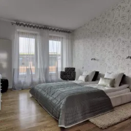 Pokój w Świdnicy z podwójnym łóżkiem, szarą pikowaną narzutą i ścianą pokrytą tapetą w kwiatowe wzory. Widoczne białe zasłony i drewniana podłoga.