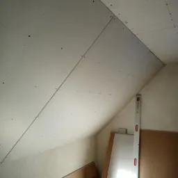 Skośny sufit poddasza w trakcie montażu płyt kartonowo-gipsowych, widoczne wkręty, poziomica oparta o ścianę i składowane płyty.