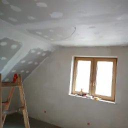 Poddasze w trakcie prac wykończeniowych: fragment ściany z nałożoną gładzią, drewniana drabina oparta o ścianę, okno z butelką i narzędziami na parapecie, sufit z widocznymi łatami gipsowymi.