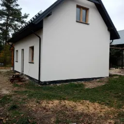 Boczny widok domu po renowacji elewacji, z widoczną białą ścianą, czarnym dachem i elementami wykończeniowymi, na trawniku rozsypane trociny.