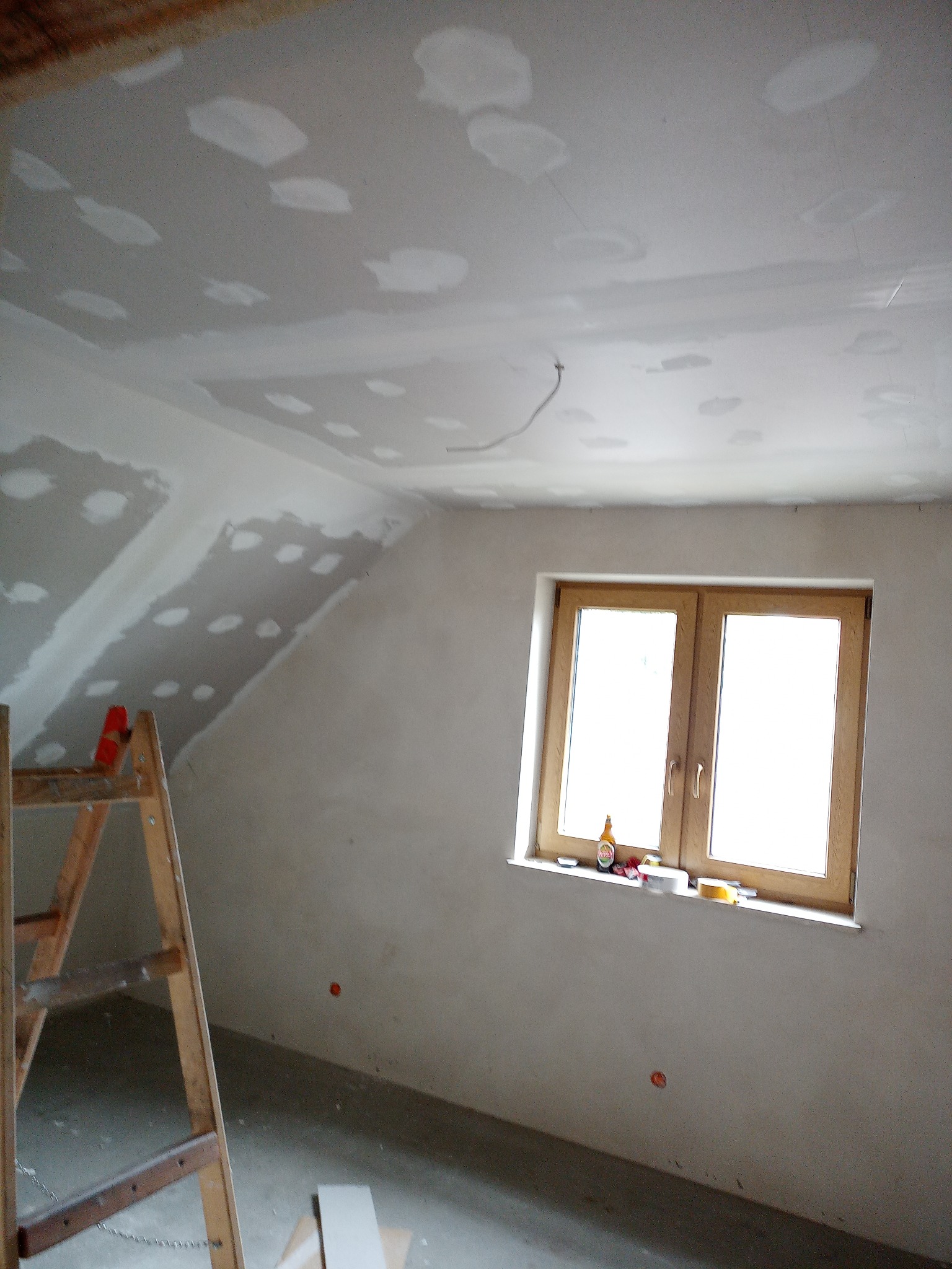 Poddasze w trakcie prac wykończeniowych: fragment ściany z nałożoną gładzią, drewniana drabina oparta o ścianę, okno z butelką i narzędziami na parapecie, sufit z widocznymi łatami gipsowymi.