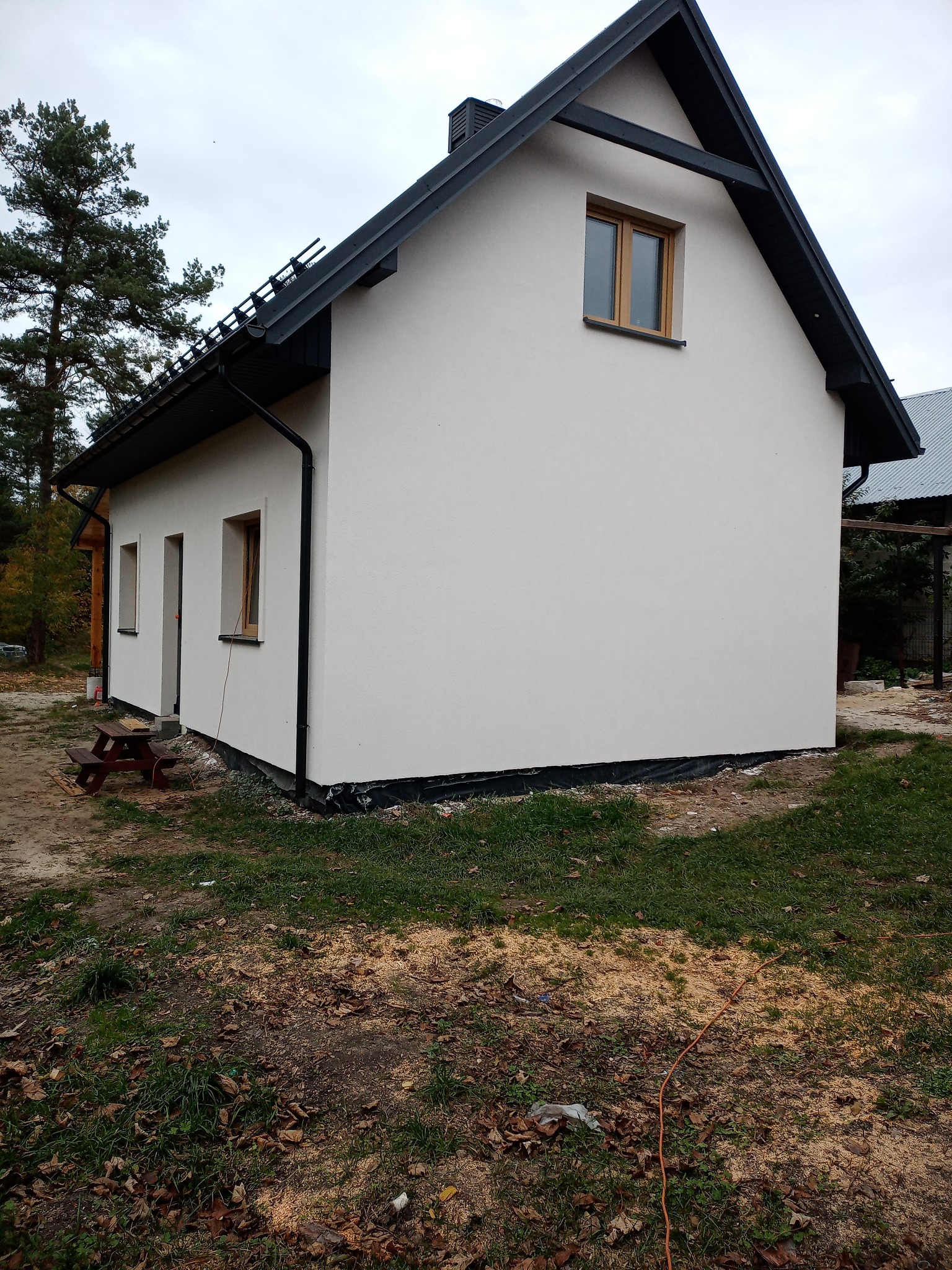 Boczny widok domu po renowacji elewacji, z widoczną białą ścianą, czarnym dachem i elementami wykończeniowymi, na trawniku rozsypane trociny.