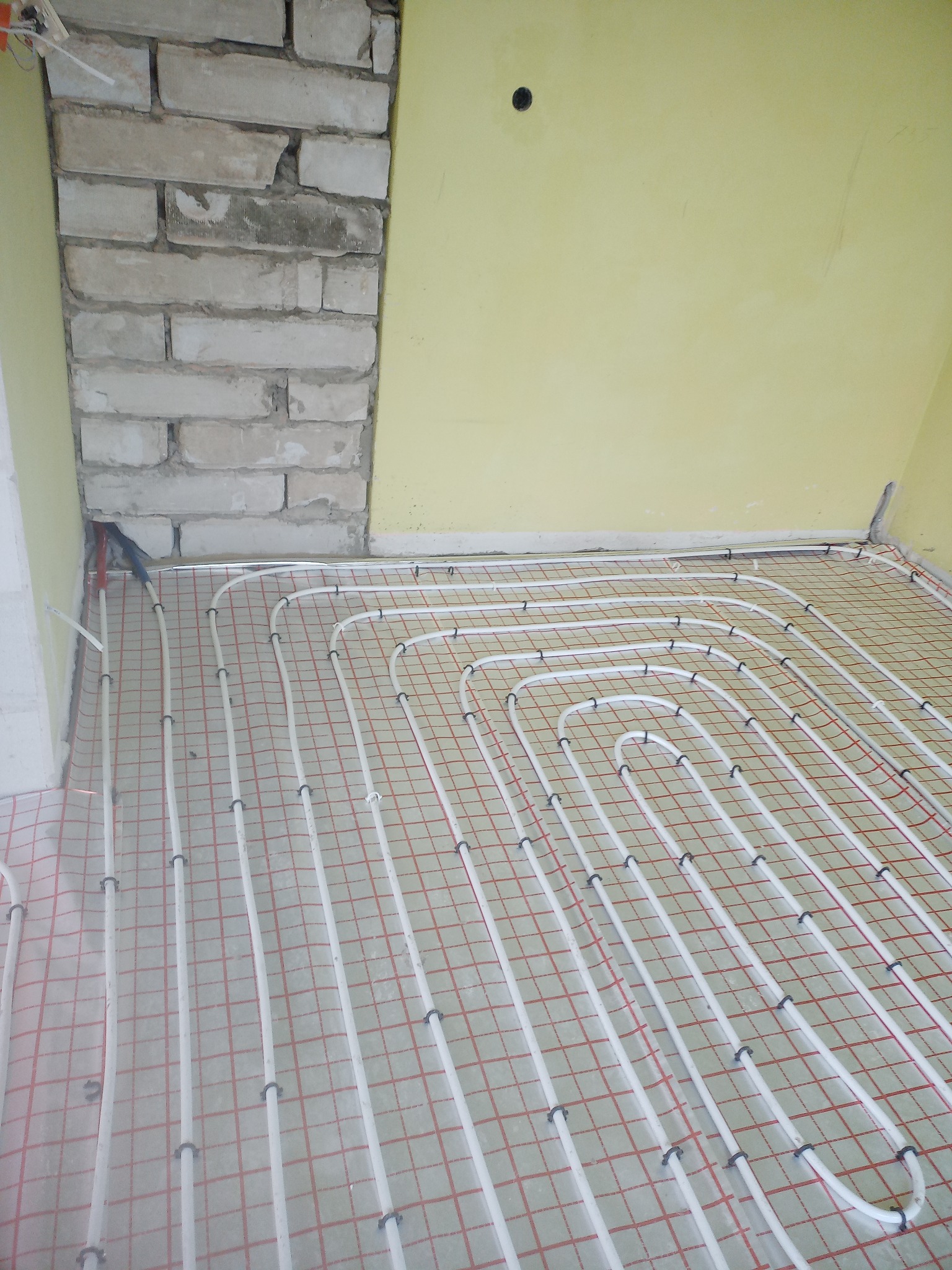 Instalacja ogrzewania podłogowego z rur w białym kolorze, przymocowanych do podłoża z czerwoną siatką, widok na fragment ściany z cegły i żółtą ścianę w tle.