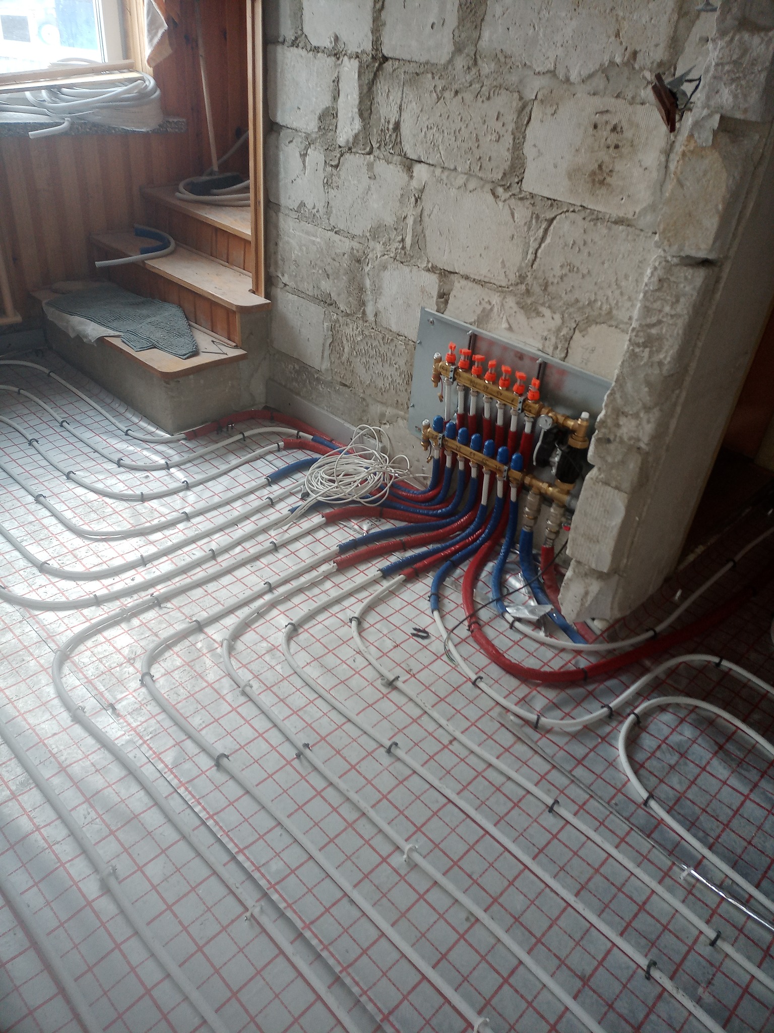 Instalacja ogrzewania podłogowego w surowym wnętrzu, widoczny rozdzielacz z rurami czerwonymi i niebieskimi, białe rury grzewcze ułożone na podłodze z siatką, fragment ściany z bloczków i drewniane...