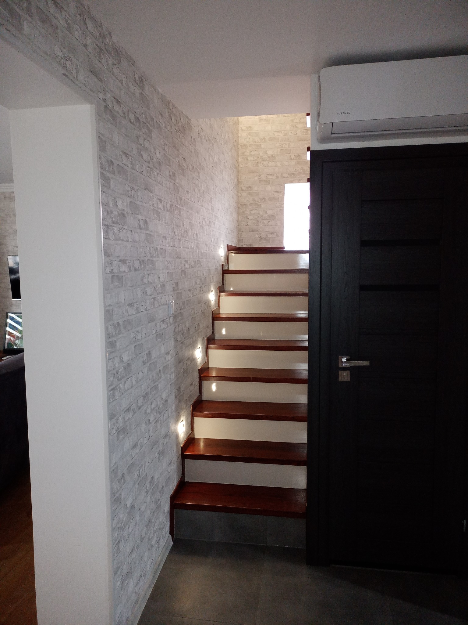 Klatka schodowa z drewnianymi stopniami i białymi podstopnicami, oświetlona małymi lampkami w ścianie z imitacji białej cegły, obok czarne drzwi i klimatyzator.