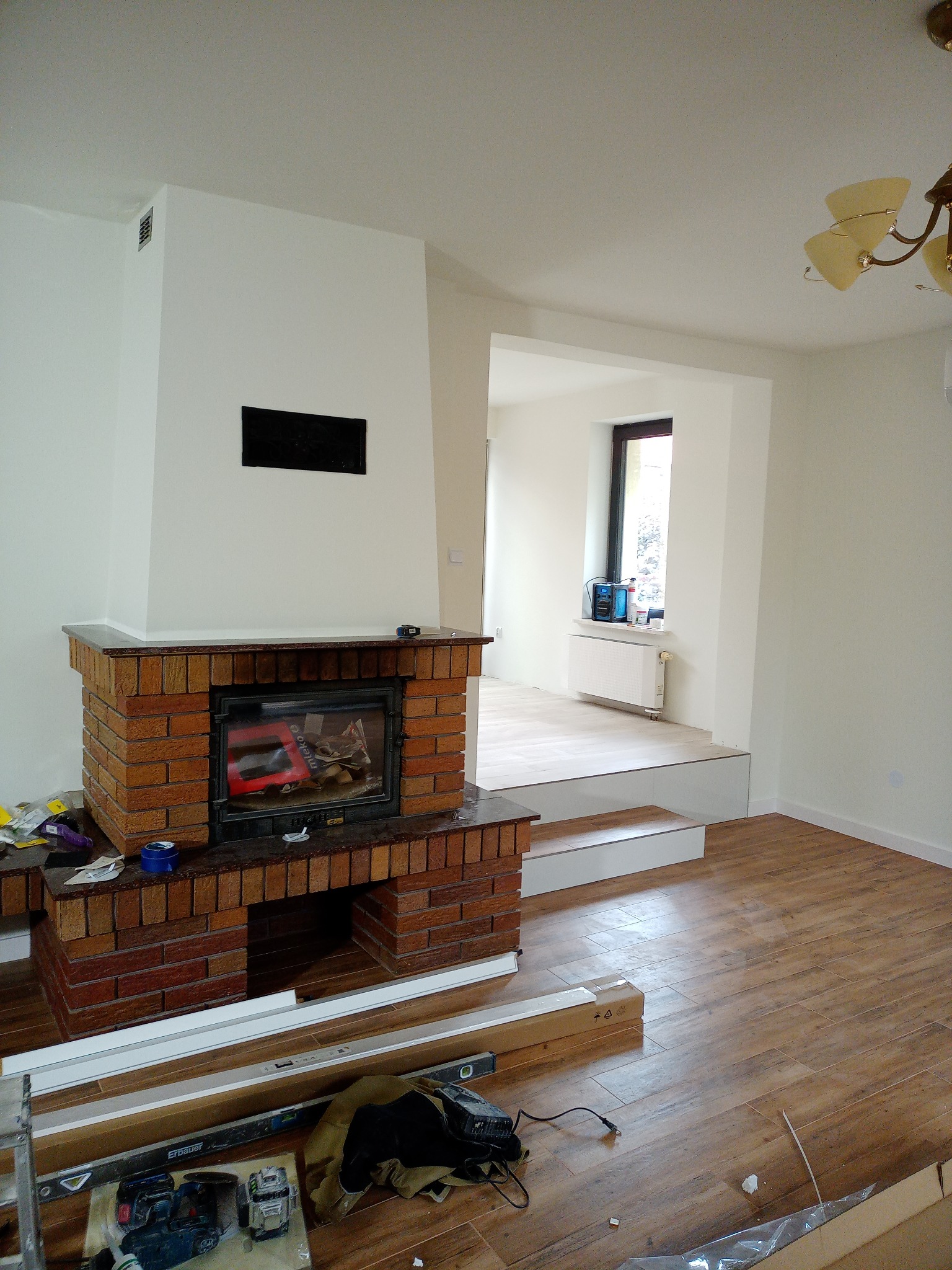 Wnętrze w trakcie remontu z widocznym kominkiem z czerwonej cegły, fragmentem drewnopodobnej podłogi, narzędziami i materiałami budowlanymi, oraz przejściem do innego pomieszczenia z oknem.
