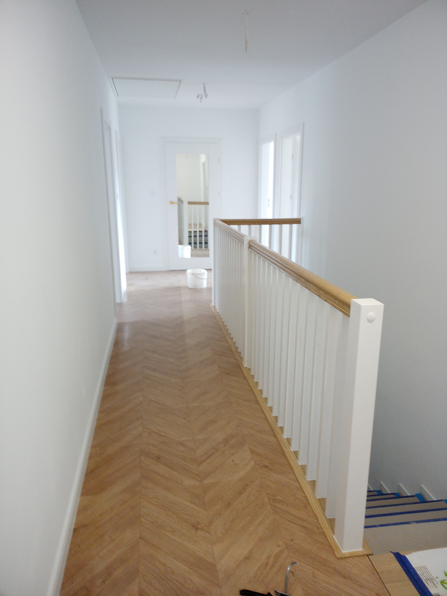Białe, świeżo pomalowane wnętrze z drewnianą podłogą ułożoną w jodełkę, białą balustradą z drewnianym poręczą i schodami zabezpieczonymi taśmą malarską.