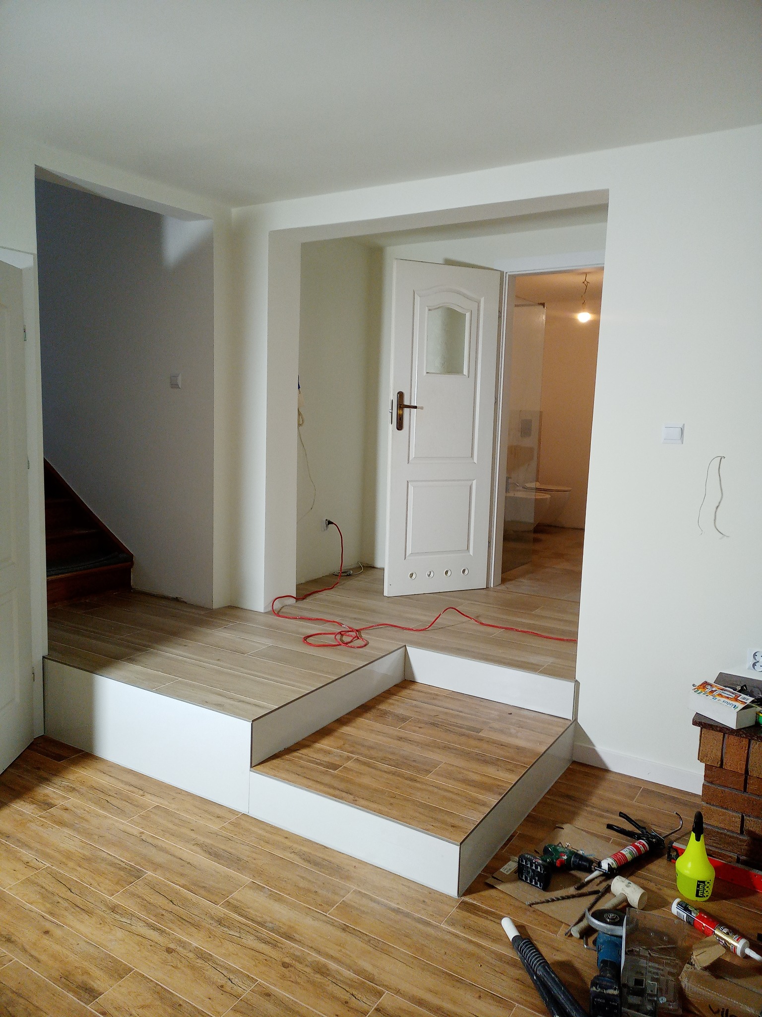 Wnętrze w trakcie prac wykończeniowych: podwyższenie podłogi z płytek imitujących drewno, białe ściany, otwarte drzwi, widoczne narzędzia i materiały budowlane na podłodze.
