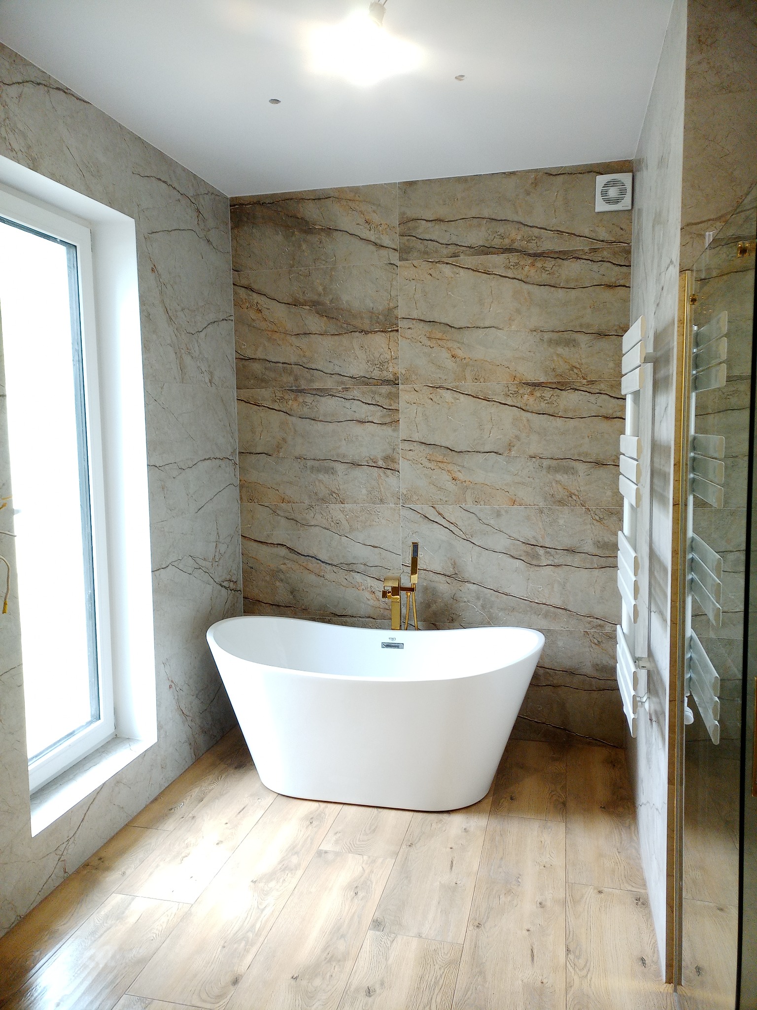 Nowoczesna łazienka z wolnostojącą białą wanną i złotą armaturą, marmurowe płytki na ścianach, drewnopodobna podłoga, grzejnik drabinkowy i wentylator w suficie.