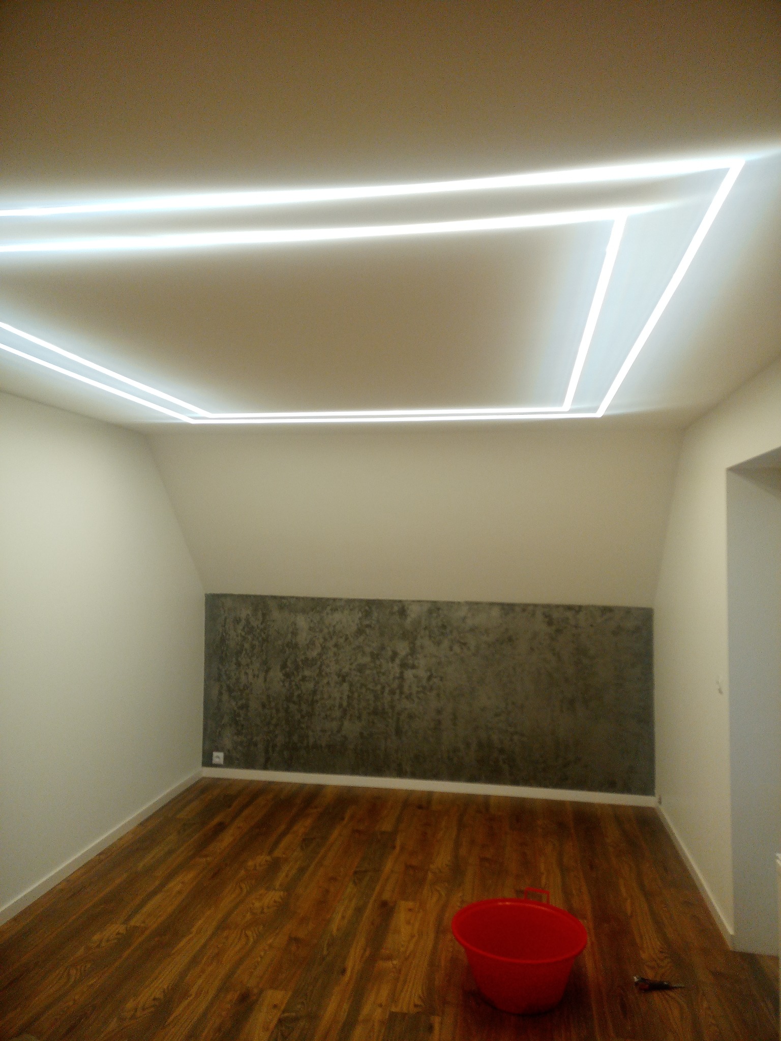 Pokój po remoncie z drewnianą podłogą, ścianą w odcieniach szarości i nowoczesnym oświetleniem sufitowym w kształcie prostokątów; czerwone wiadro stoi na podłodze.