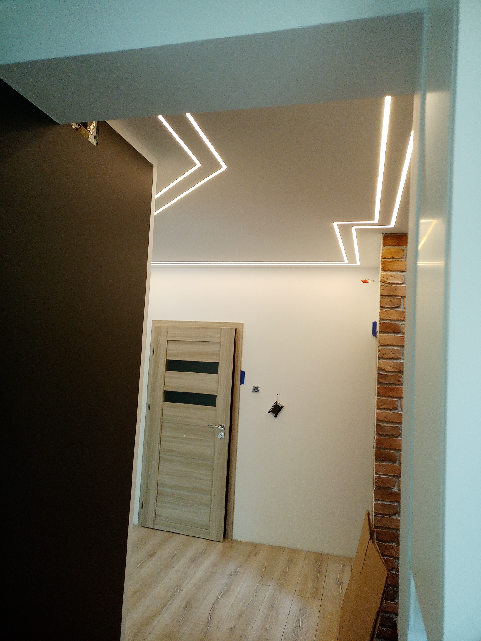 Korytarz z nowoczesnym oświetleniem LED w kształcie zygzaków na suficie, drewnianą podłogą i drzwiami z ciemnymi paskami, wykończony cegłą klinkierową.