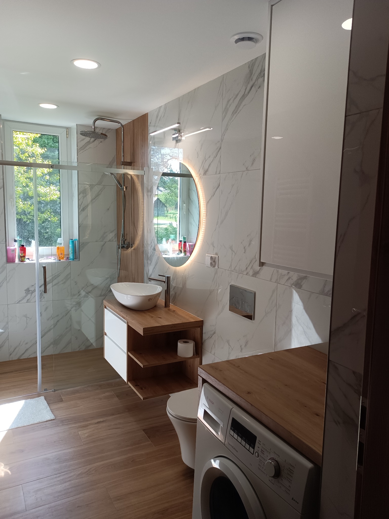 Nowoczesna łazienka z marmurowymi płytkami, kabiną prysznicową typu walk-in, okrągłym lustrem z podświetleniem LED i drewnianymi akcentami. Widoczna pralka z blatem.