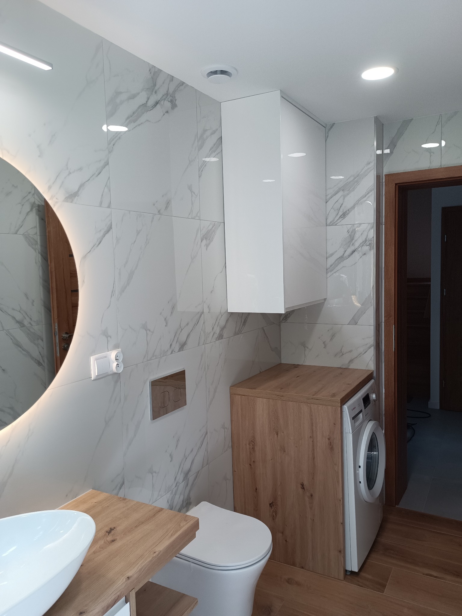 Nowoczesna łazienka z marmurowymi płytkami, okrągłym lustrem z podświetleniem LED, drewnianym blatem i zabudowaną pralką. Minimalistyczny design, jasne oświetlenie.
