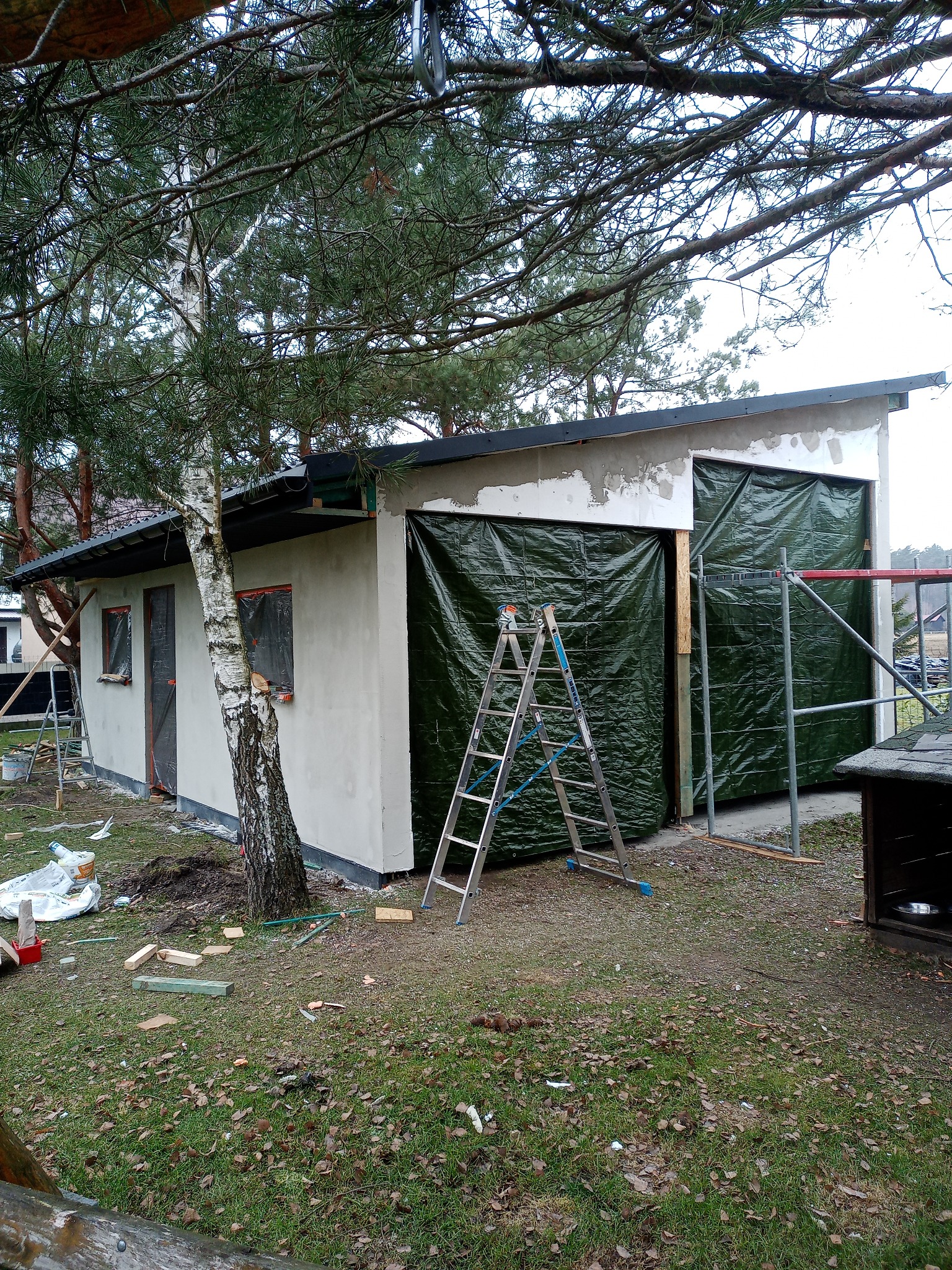 Ocieplony budynek gospodarczy w trakcie tynkowania, zabezpieczony folią, z drabiną i rusztowaniem na zewnątrz. Widoczne narzędzia i materiały budowlane.