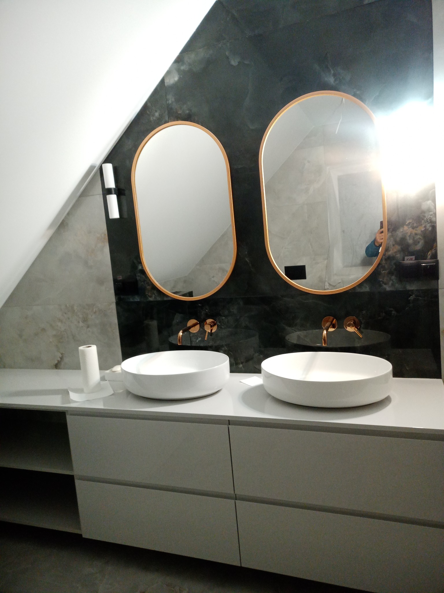 Nowoczesna łazienka na poddaszu z dwoma owalnymi umywalkami nablatowymi, złotą armaturą i lustrami w złotej ramie na tle ściany z płytek imitujących marmur.