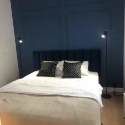 Sypialnia z ciemnoniebieską ścianą z dekoracyjnymi panelami, oświetlona dwoma lampami podłogowymi, z łóżkiem z szarą narzutą i wzorzystymi poduszkami.