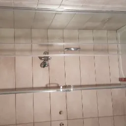 Zamontowana szklana kabina prysznicowa z widoczną baterią, poziomnicą i narzędziami wewnątrz brodzika, na tle beżowych płytek ściennych.