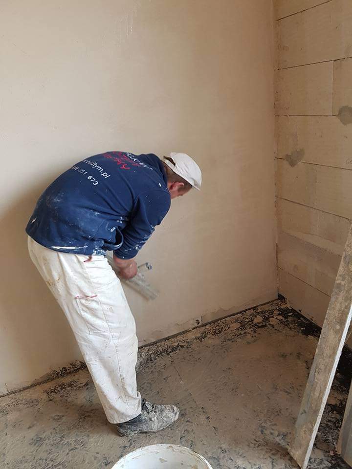 Pracownik w ubrudzonych roboczych spodniach nakłada gładź szpachlową na ścianę w narożniku pomieszczenia, widoczna ściana z bloczków betonu komórkowego i wiadro z zaprawą.