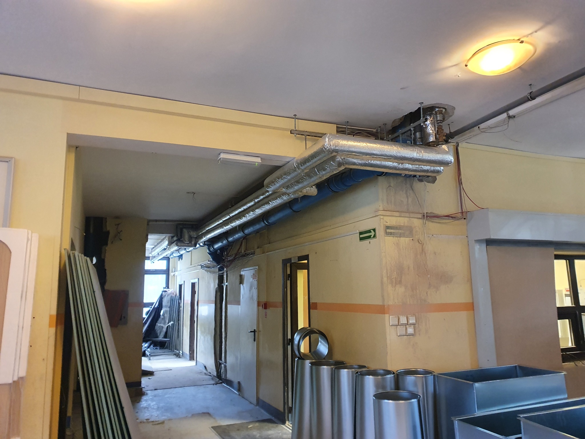 Korytarz w trakcie remontu z widocznymi rurami wentylacyjnymi pod sufitem, materiałami budowlanymi i elementami instalacji wentylacyjnej na podłodze.