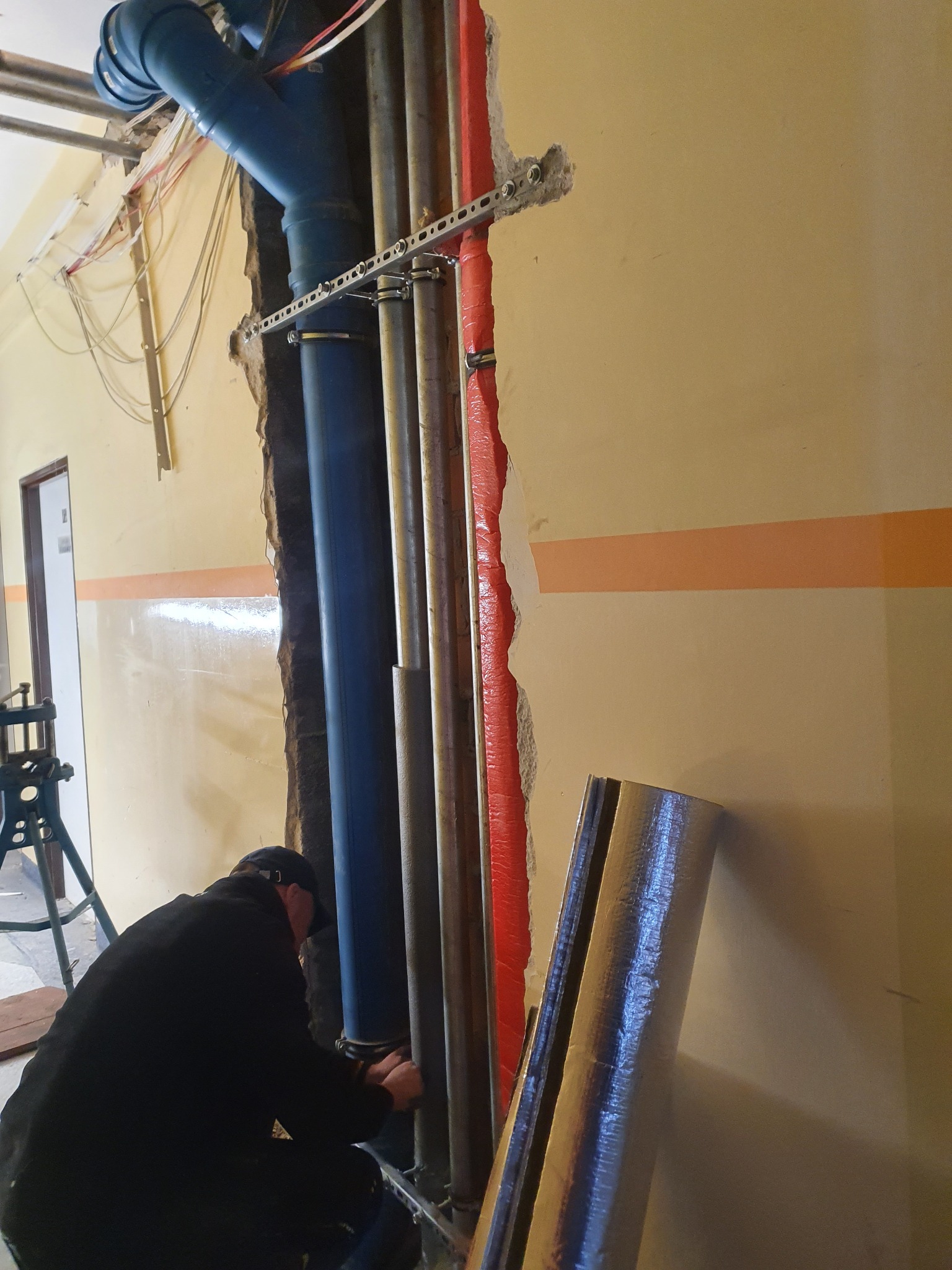 Instalator mocuje rury do ściany z widoczną izolacją termiczną, w tle fragment pomieszczenia z pomalowaną ścianą i drzwiami.