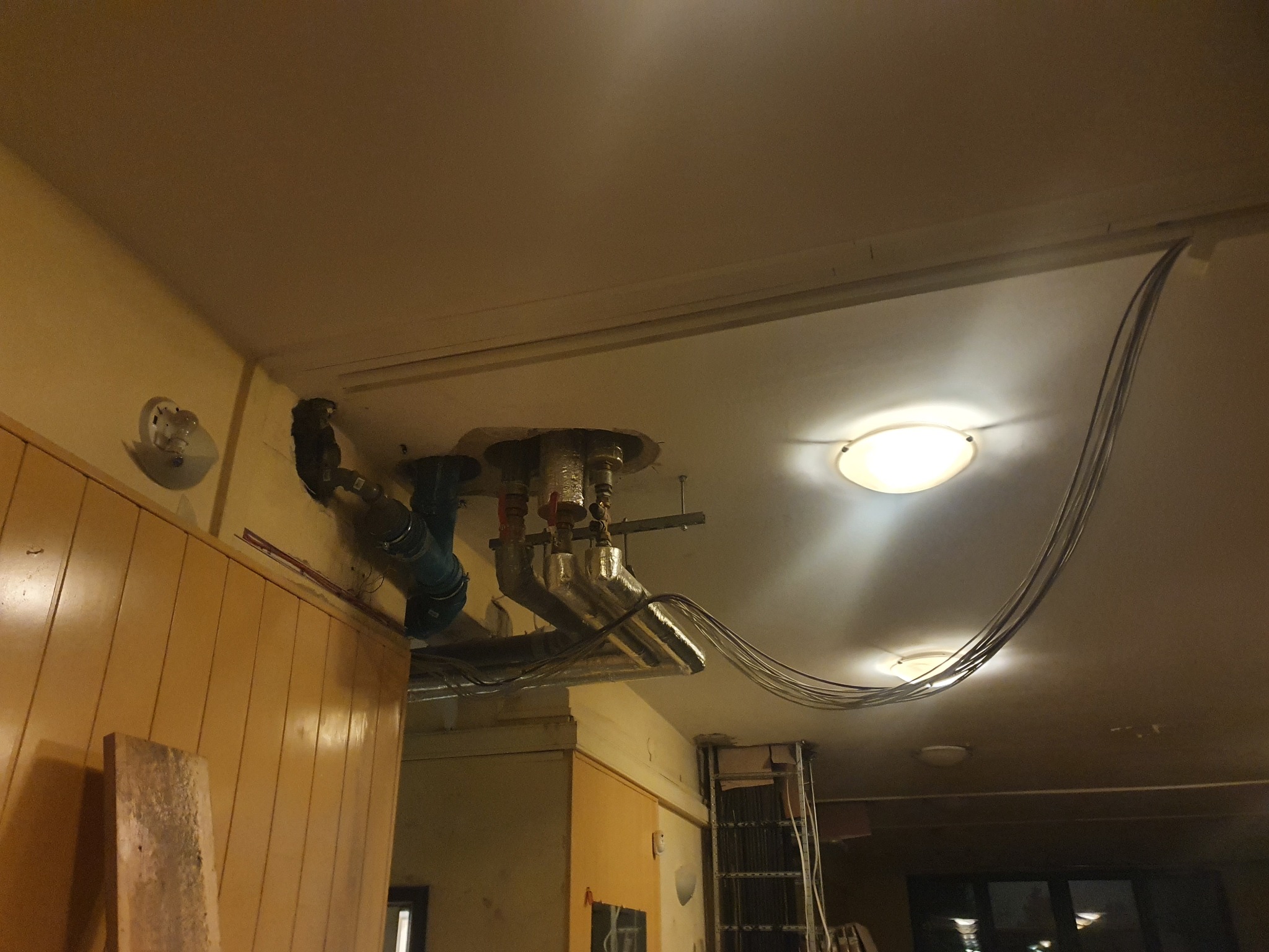 Widok instalacji pod sufitem, rury i przewody w trakcie montażu, widoczne elementy izolacji i podłączenia, oświetlenie sufitowe w tle.