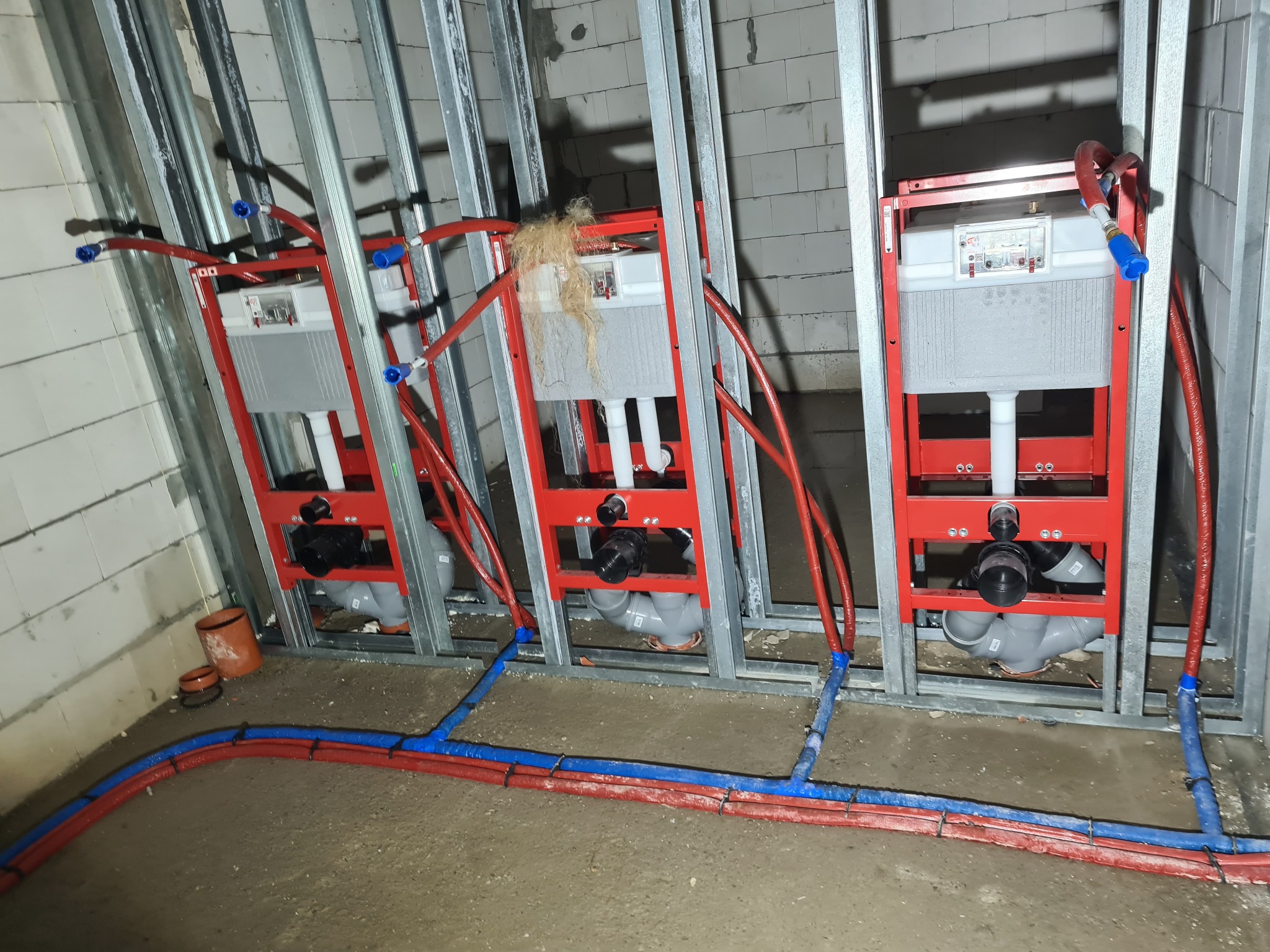 Trzy czerwone stelaże podtynkowe WC zamontowane w metalowej konstrukcji szkieletowej, z podłączonymi rurami w kolorze czerwonym i niebieskim, widoczne elementy instalacji kanalizacyjnej i wodnej.