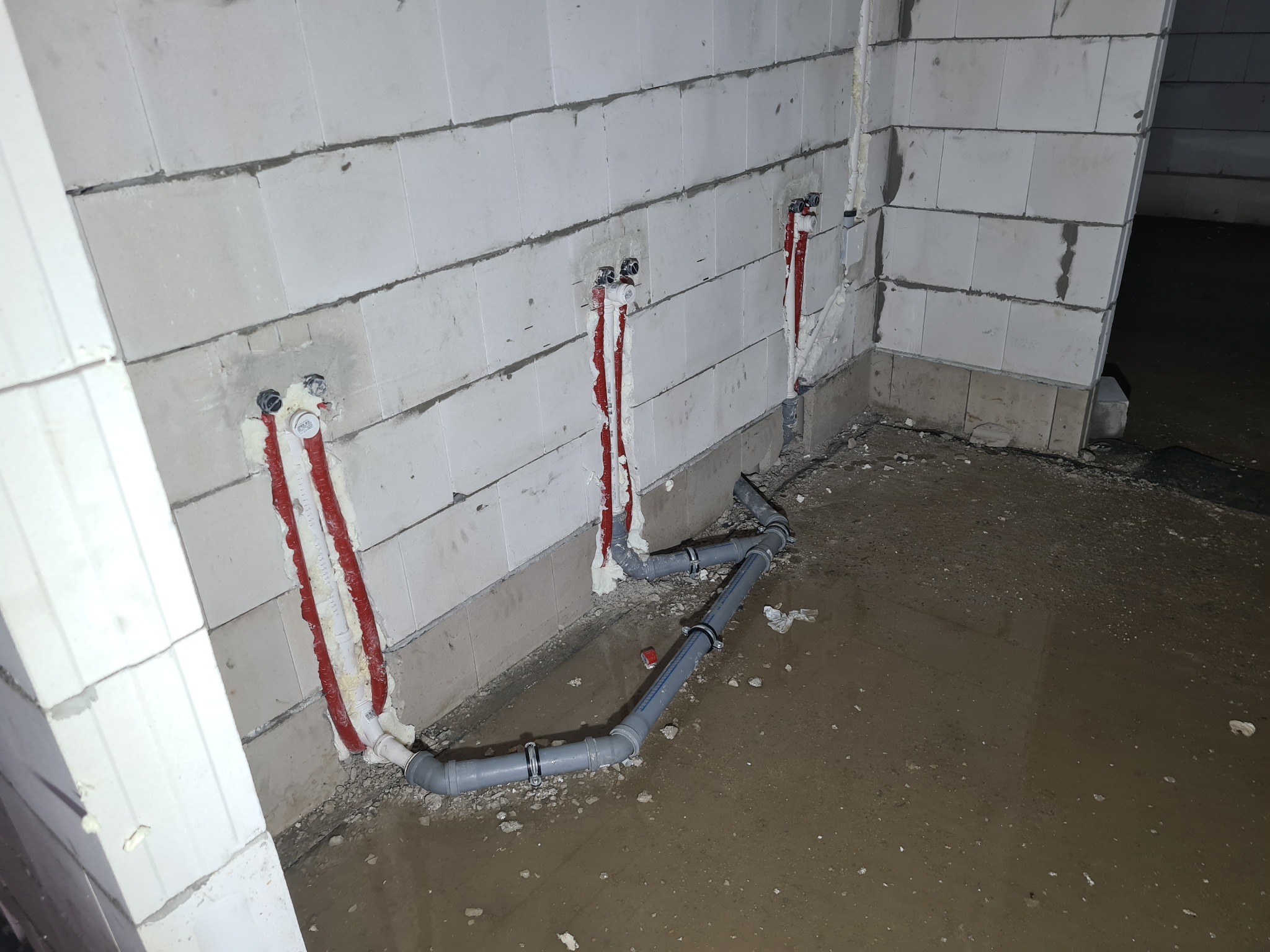 Surowe wnętrze w trakcie prac hydraulicznych: rury kanalizacyjne z obejmami na mokrej podłodze, pionowe rury instalacyjne w ścianie z betonu komórkowego zabezpieczone pianką montażową.