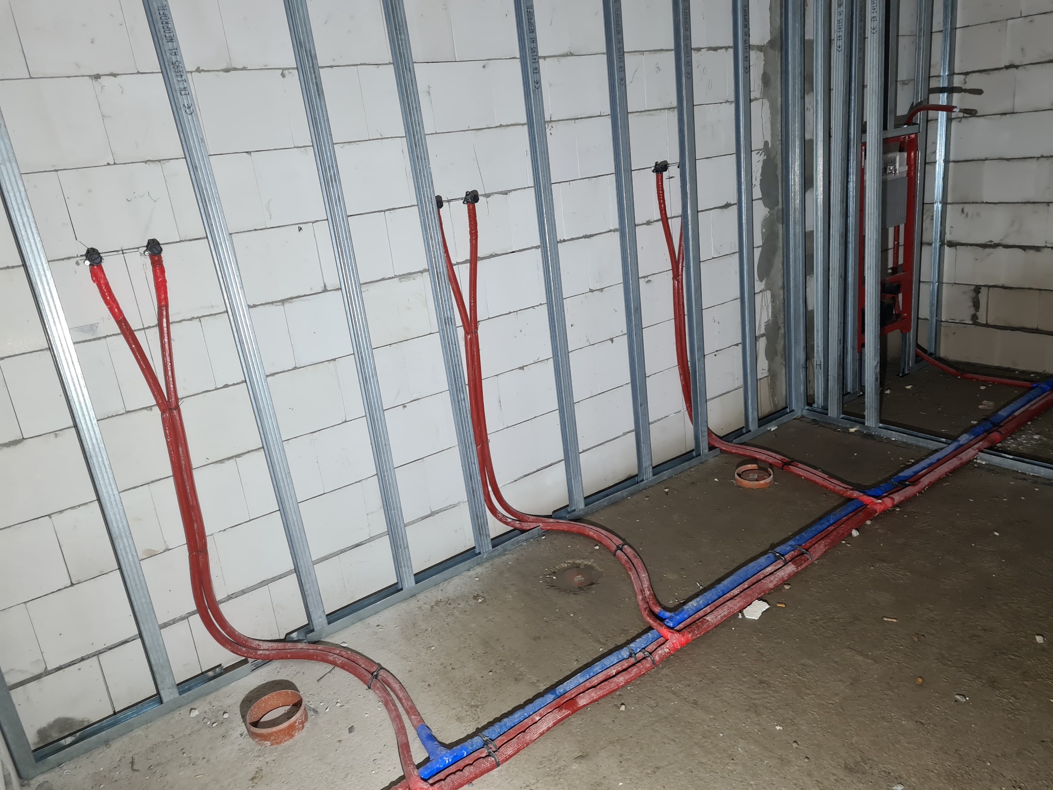 Instalacja wodna podtynkowa w trakcie montażu: czerwone i niebieskie rury prowadzone wzdłuż metalowego stelażu przy ścianie z białych bloczków, widoczne złącza i fragment instalacji podtynkowych...