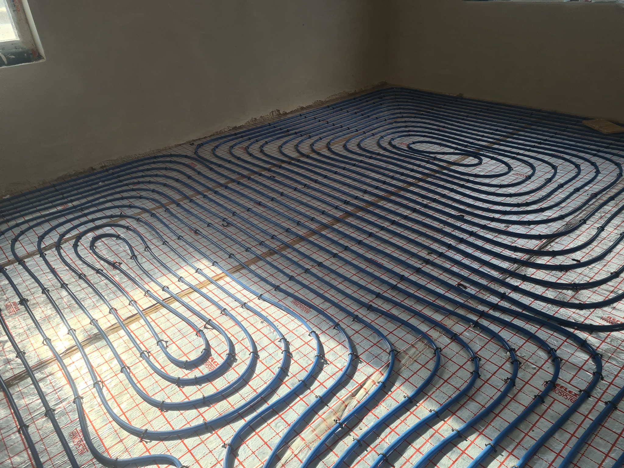 Układanie niebieskich rur ogrzewania podłogowego na folii z czerwoną siatką w surowym wnętrzu, widoczne światło słoneczne padające przez okno.