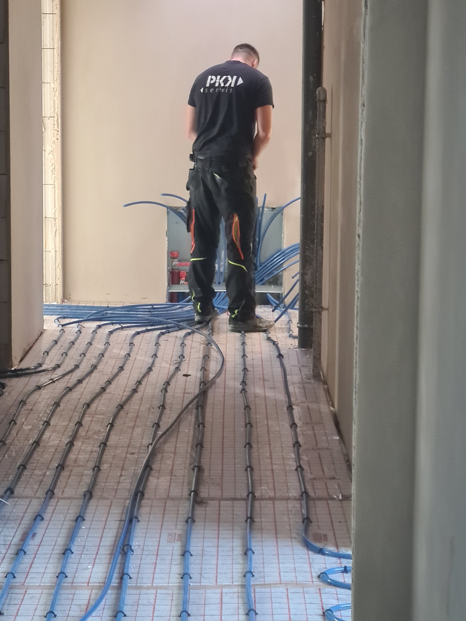 Instalacja ogrzewania podłogowego: niebieskie rurki układane na izolacji, widoczny instalator w czarnej koszulce z logo firmy, przygotowujący się do podłączenia instalacji.