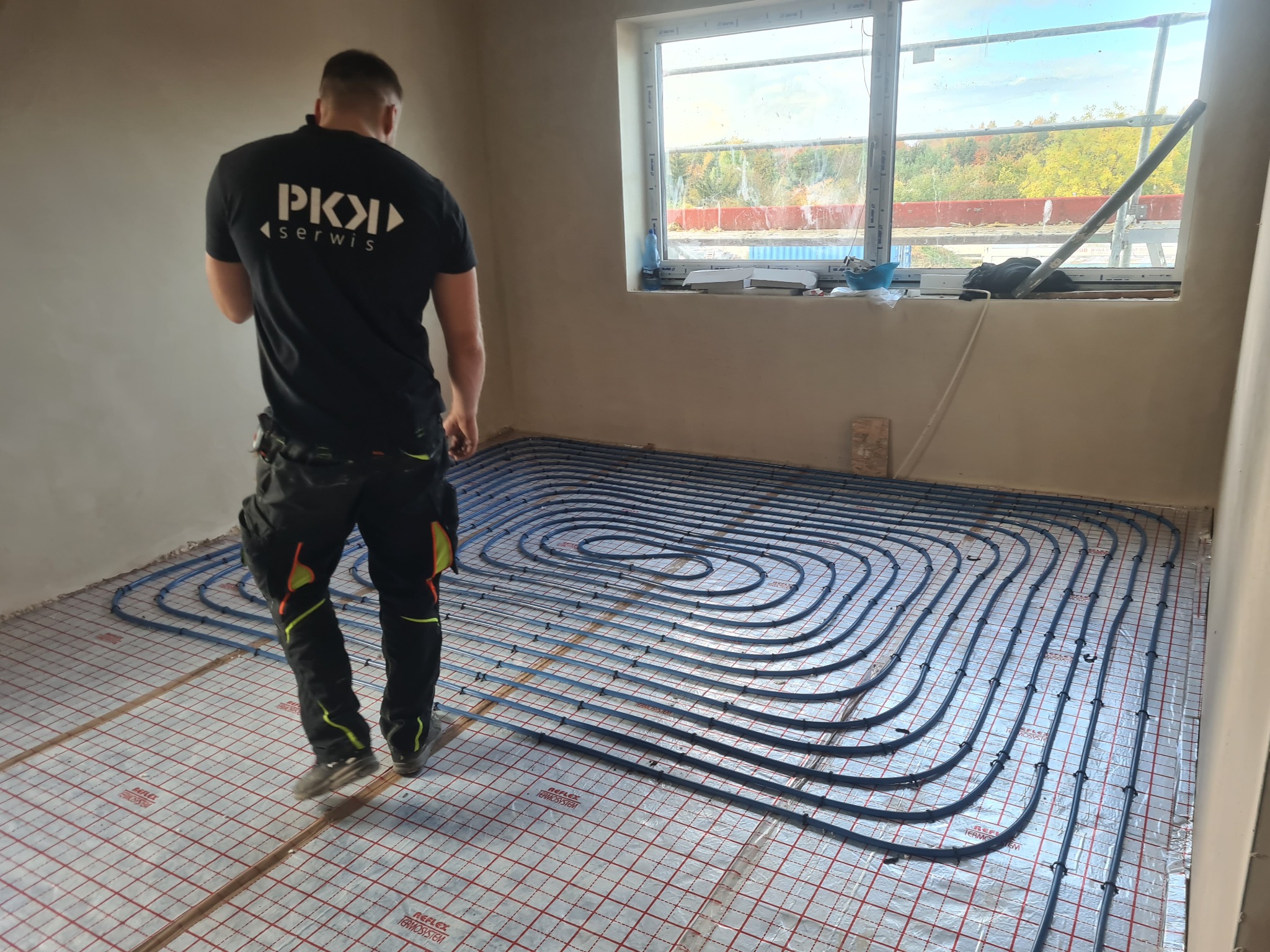 Instalacja ogrzewania podłogowego w nowym budynku; widoczny pracownik w firmowej koszulce z logo PKI Serwis oraz pętle rur grzewczych ułożone na folii refleksyjnej z siatką, okno w tle z widokiem...