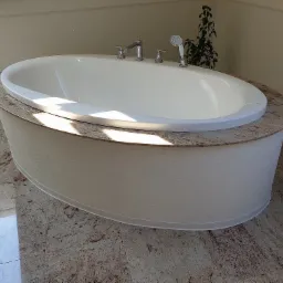 Owalna wanna obudowana jasnym, teksturowanym panelem z blatem z polerowanego kamienia na podłodze z płytek imitujących kamień.