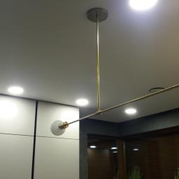 Rinam - Nowoczesna, złota lampa sufitowa z dwoma kulistymi kloszami, zamontowana na tle białych szafek i drewnianej ściany.