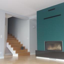 Rinam - Nowoczesny salon z drewnianymi schodami, zieloną ścianą z wbudowanym kominkiem i półkami, oraz szarą ścianą z osprzętem elektrycznym.