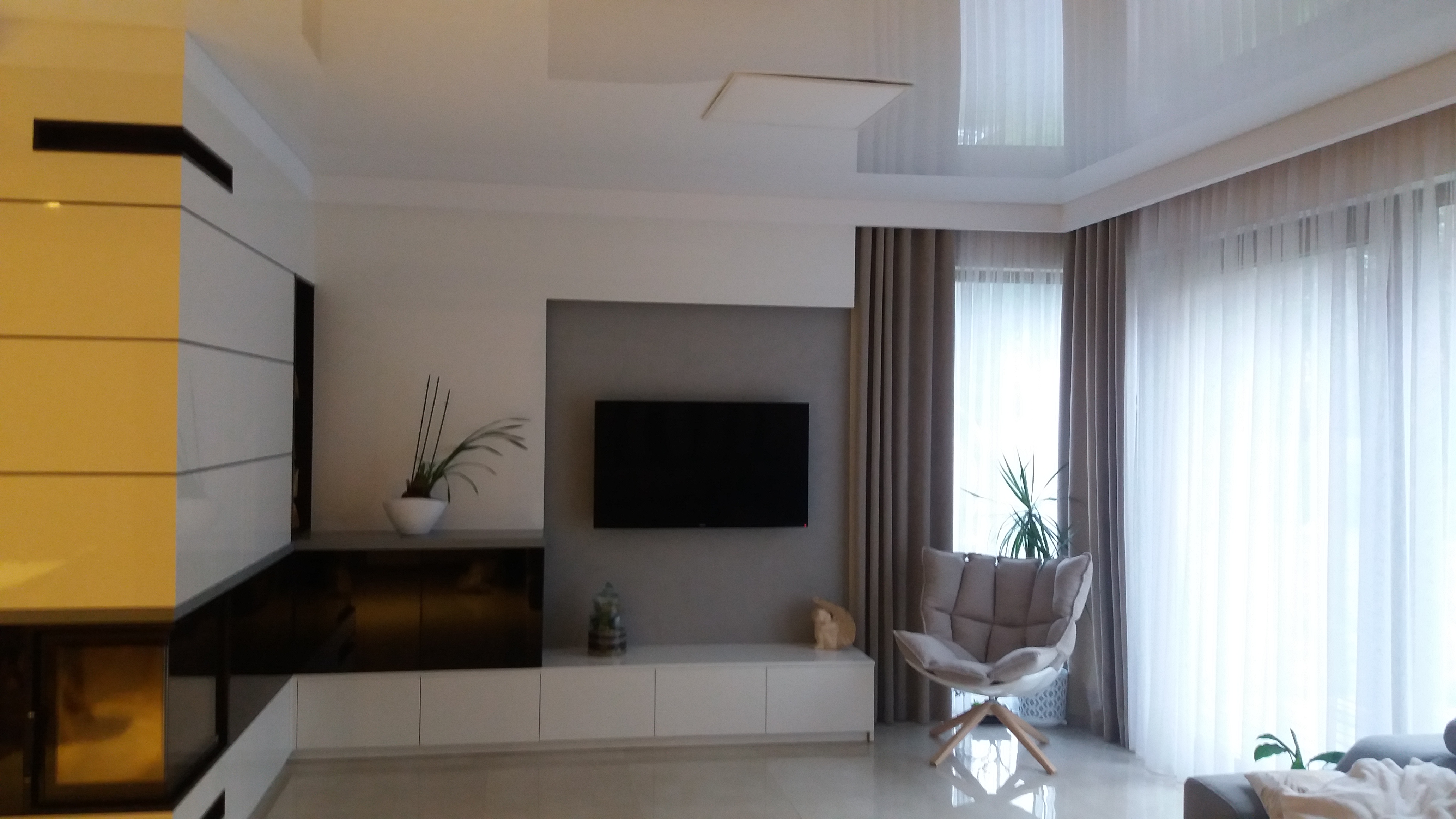 Nowoczesny salon z błyszczącym sufitem napinanym, minimalistyczną zabudową meblową z kominkiem, szarą ścianą z telewizorem i designerskim fotelem przy oknie z zasłonami.