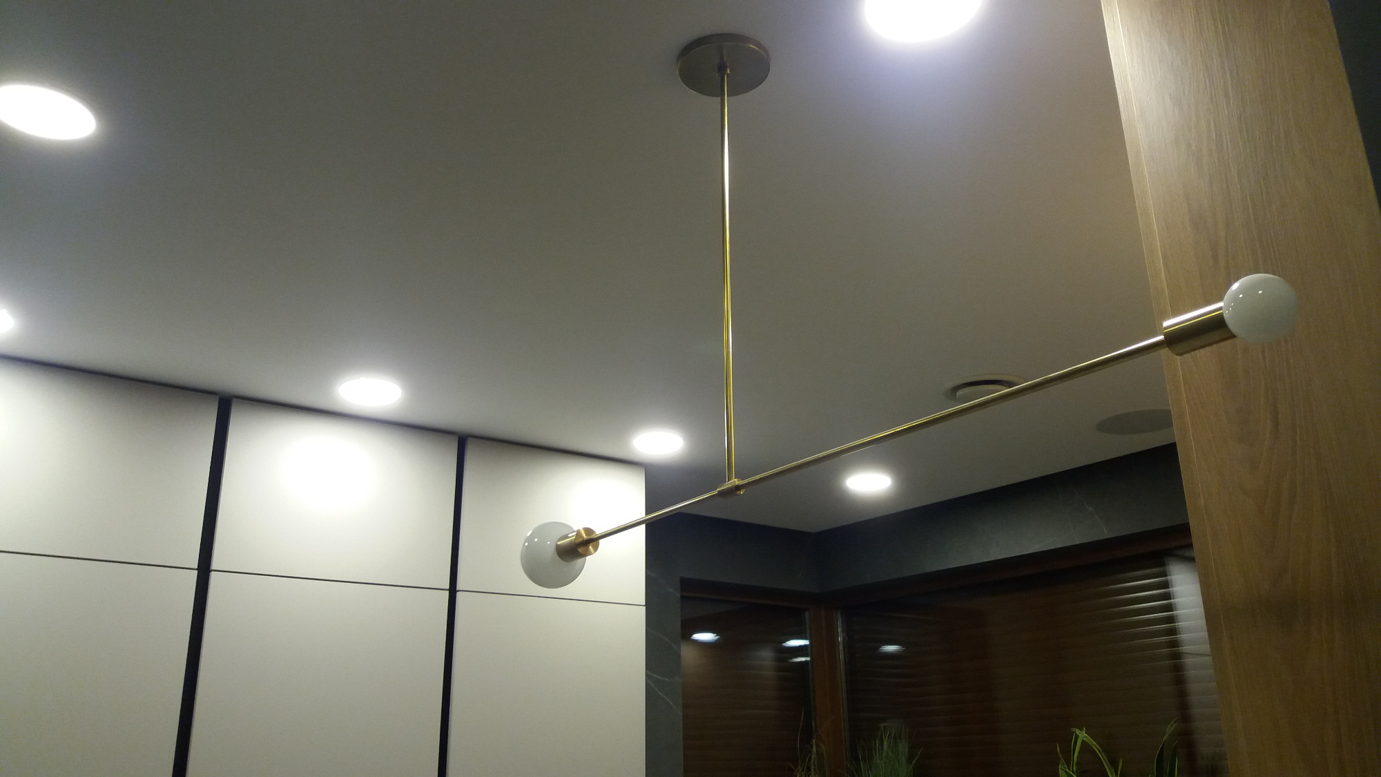 Nowoczesna, złota lampa sufitowa z dwoma kulistymi kloszami, zamontowana na tle białych szafek i drewnianej ściany.