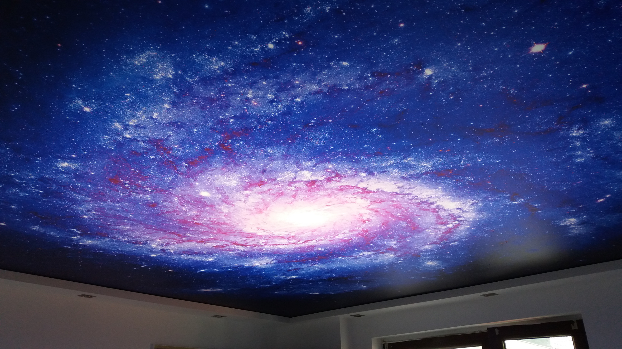 Sufit napinany z nadrukiem galaktyki spiralnej w odcieniach niebieskiego i różowego, widoczne halogenowe punkty oświetleniowe.