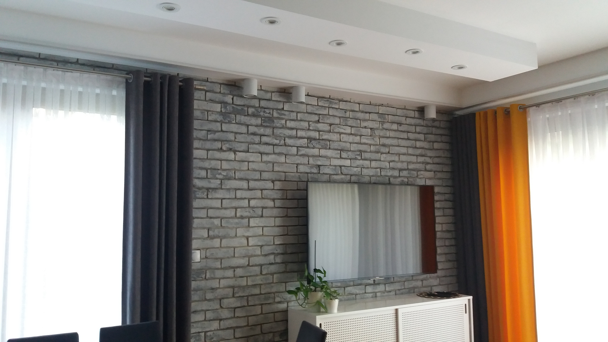Szara ściana z cegły dekoracyjnej w salonie, podświetlona reflektorami sufitowymi, z telewizorem i szafką RTV, zasłony w kolorach szarym i pomarańczowym.