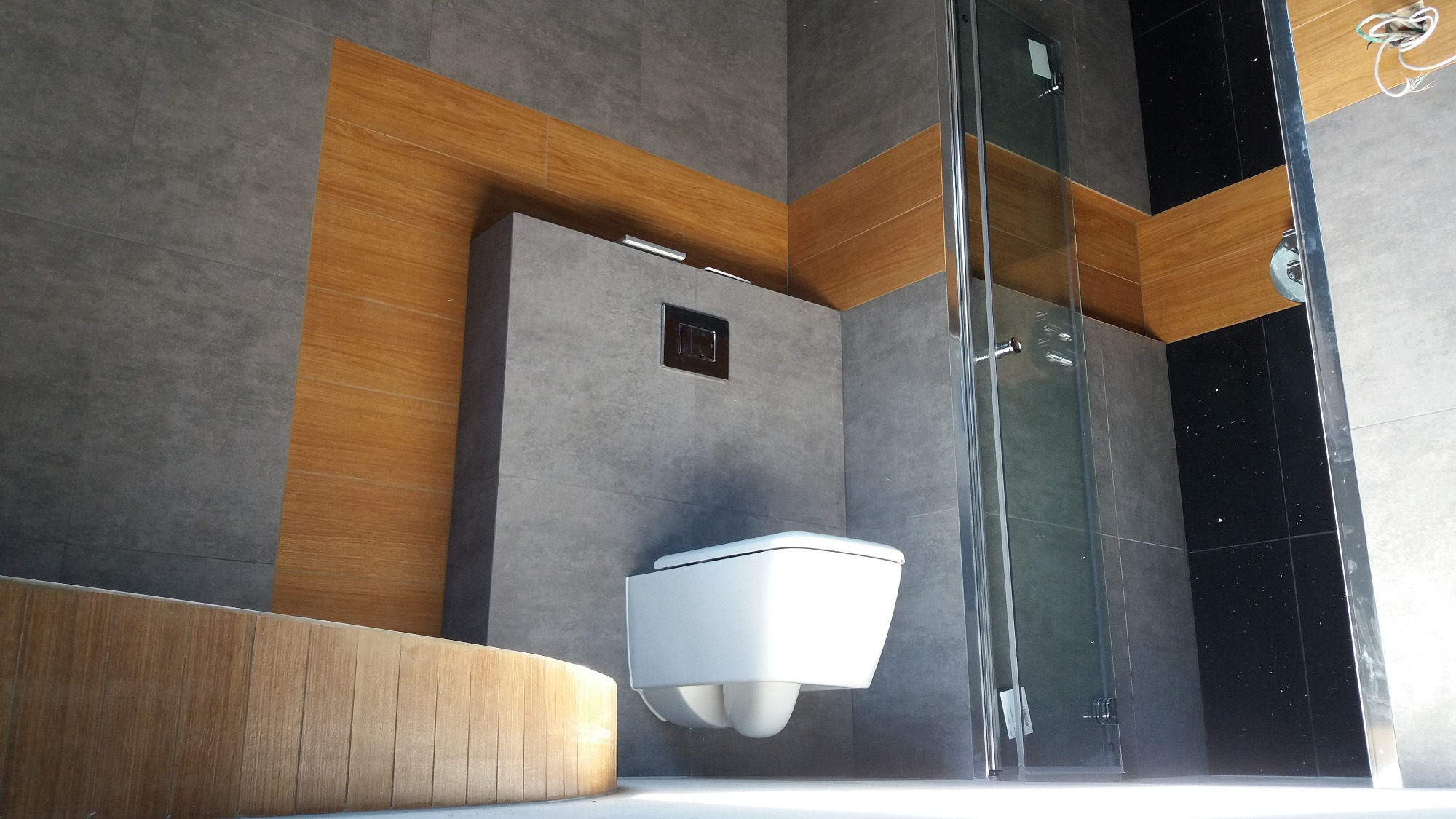 Nowoczesna łazienka z szarymi płytkami, drewnianymi akcentami i podwieszaną toaletą, widok z dołu na kabinę prysznicową.