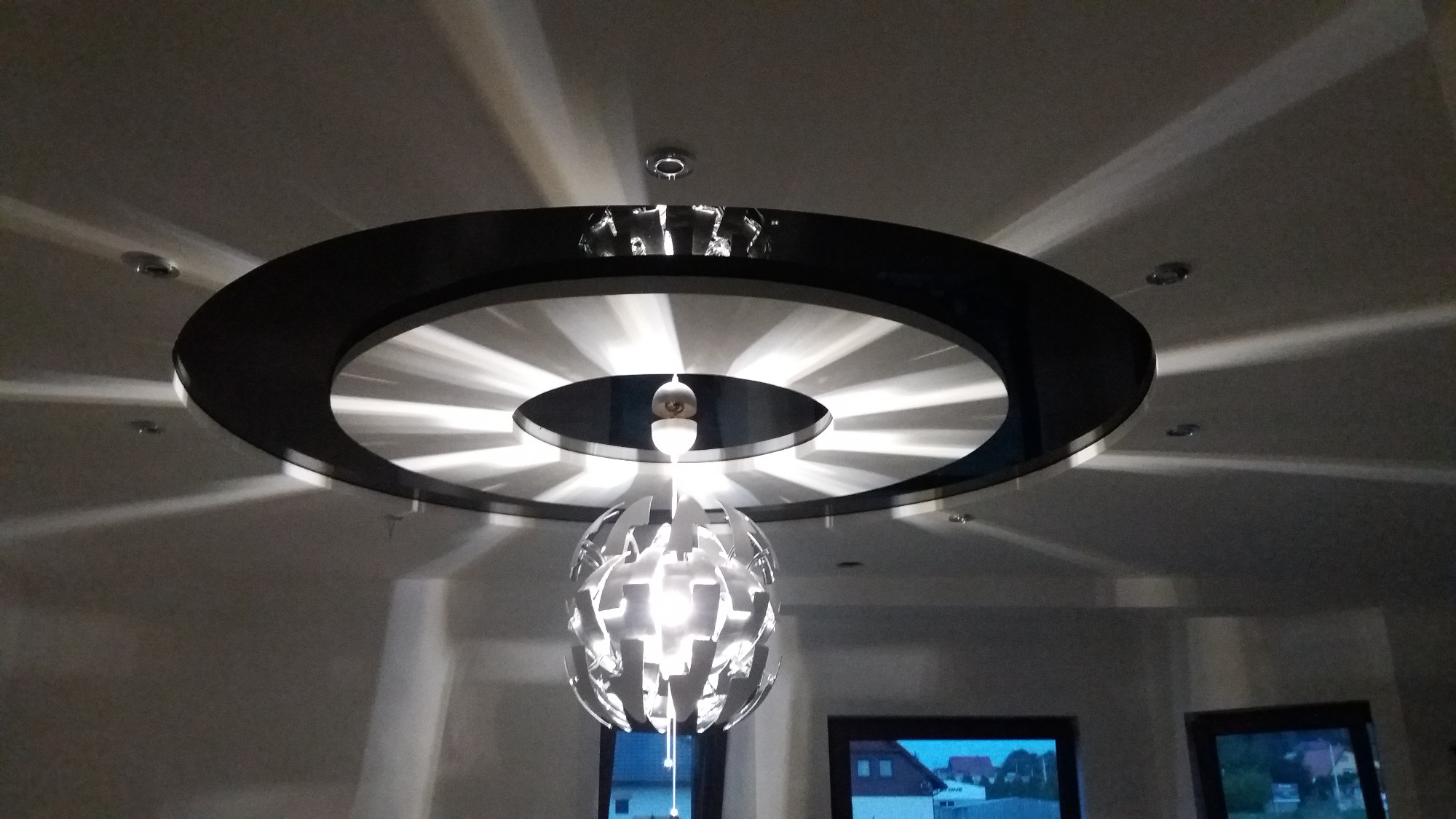 Nowoczesny sufit podwieszany z okrągłym czarnym obramowaniem i dekoracyjnym oświetleniem punktowym oraz wiszącą lampą o ażurowej konstrukcji.