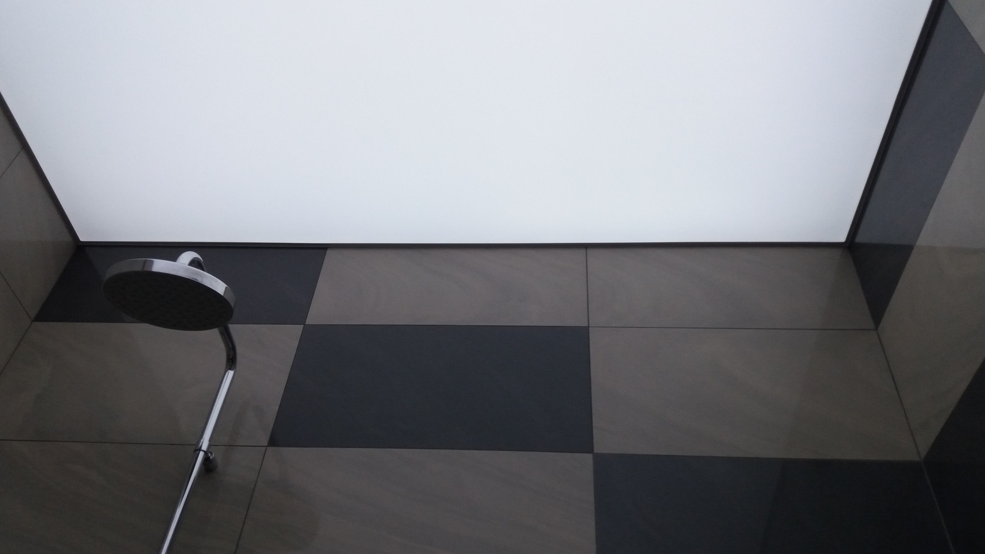 Nowoczesny prysznic z chromowaną deszczownicą, czarno-szare kwadratowe płytki na ścianie i podświetlany sufit napinany.