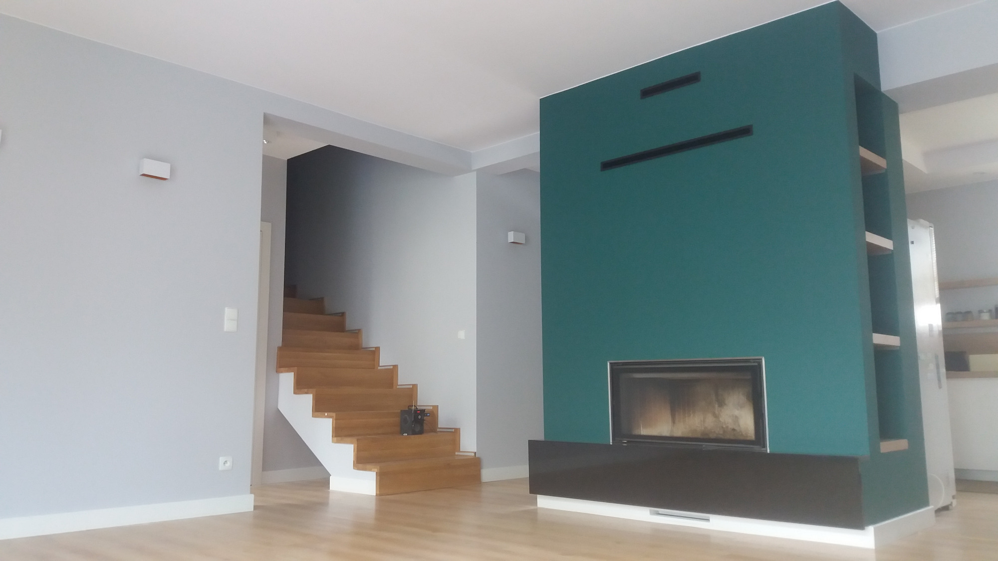 Nowoczesny salon z drewnianymi schodami, zieloną ścianą z wbudowanym kominkiem i półkami, oraz szarą ścianą z osprzętem elektrycznym.